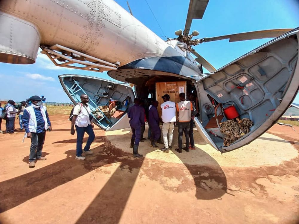 Kolme ihmistä nostaa suurta laatikkoa ulos helikopterista. Ympärillä on myös muita ihmisiä.